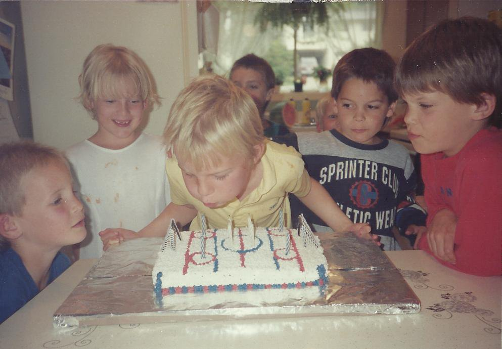 A kid in a yellow shirt blowing out the candles on a hockey rink birthday cake, crowded in by five other kids.