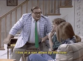 A man in a rumpled suit leaning over a sofa, captioned: I live in a van down by the river.